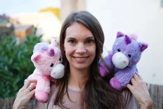 Freya, eine Frau mit langen dunklen Haaren, lächelt draußen vor Grün. Auf ihren Schultern sitzen ein rosa und ein lila Plüsch-Einhorn.